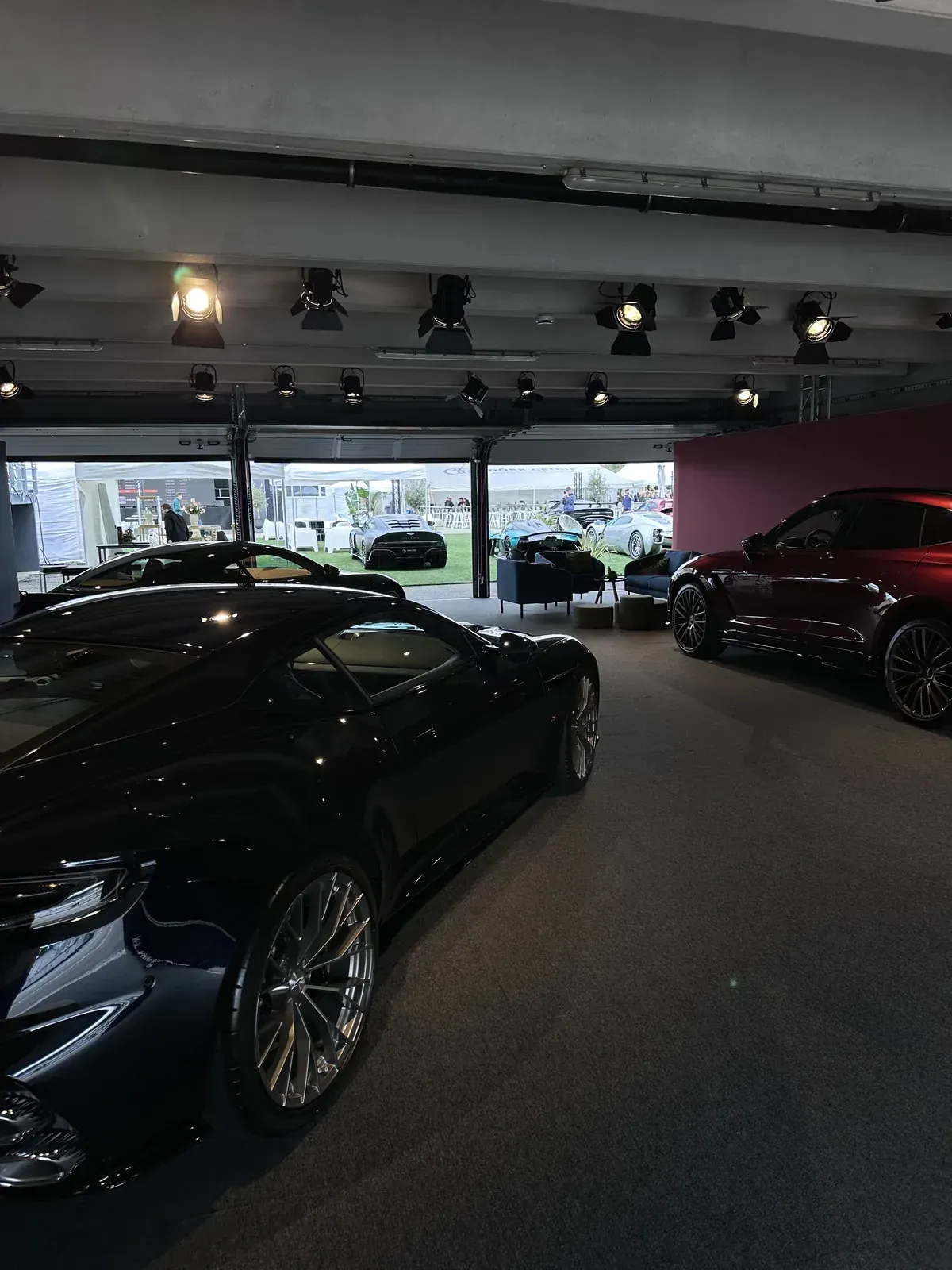 Dunkelblauer Sportwagen und rotes SUV in einem Showroom, durch das offene Tor Gäste auf einem Event-Gelände