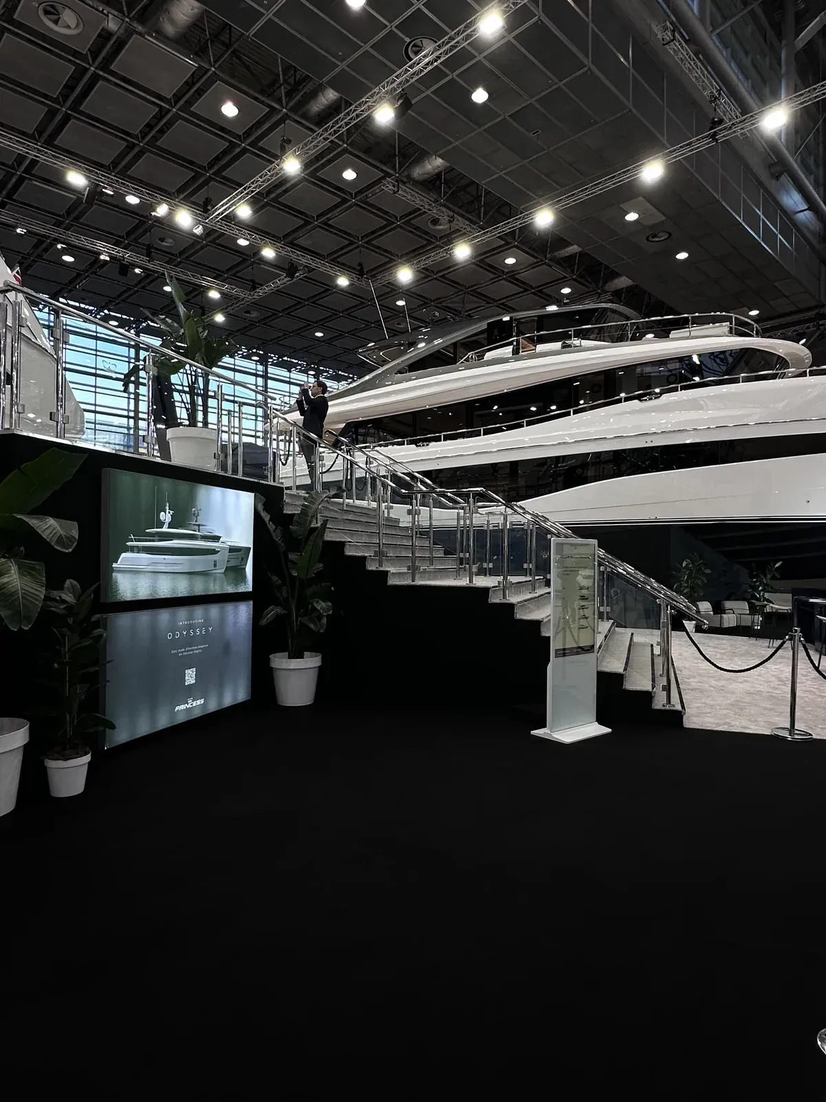 Messestand mit einer großen weißen Motoryacht auf einer Bühne, Treppe und Präsentationsbildschirm in einer Messehalle