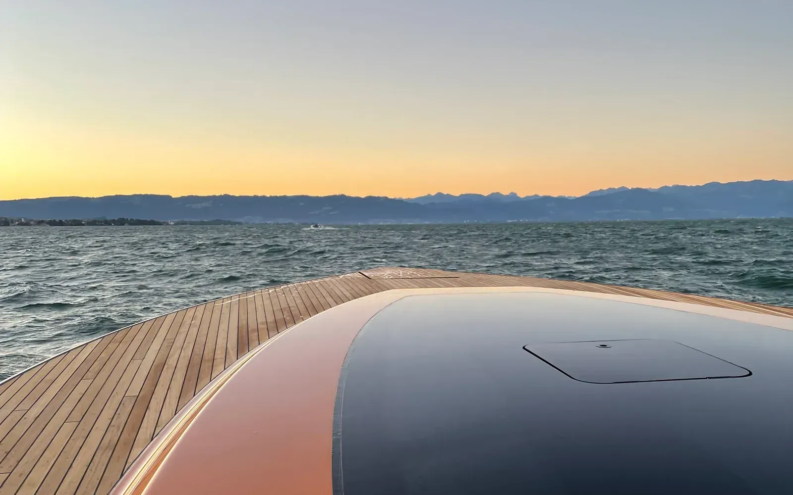 Blick über den Bug einer Motoryacht mit Teakdeck auf See und Bergpanorama im Abendlicht